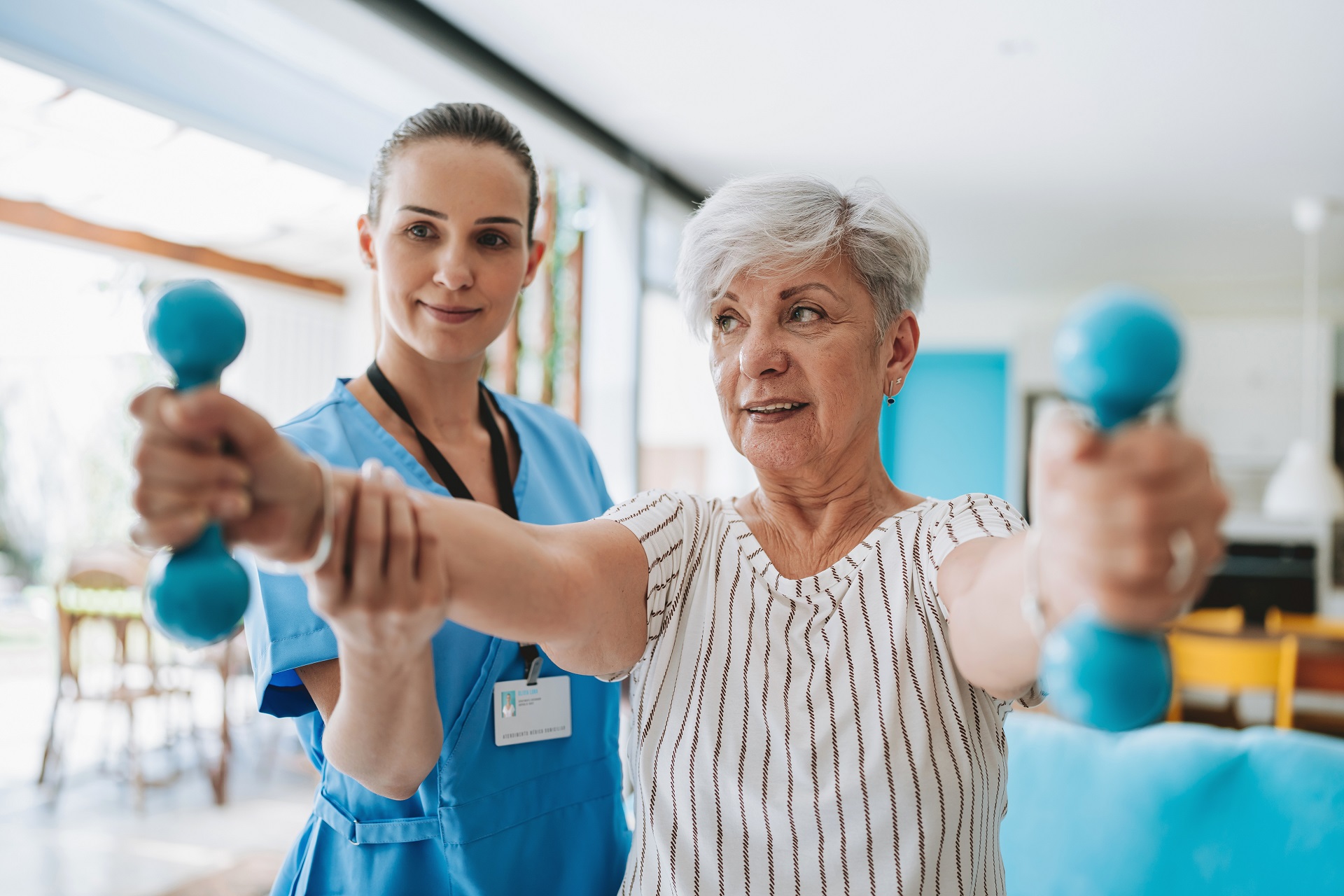 Im Vordergrund sieht man eine ältere Dame. Sie hat weißes Haar und hält in ihren Händen zwei kleine Gewichte. Hinter ihr steht eine junge Therapeutin, die die Frau bei ihrer Physiotherapie unterstützt. Im Vordergrund sieht man eine ältere Dame. Sie hat weißes Haar und hält in ihren Händen zwei kleine Gewichte. Hinter ihr steht eine junge Therapeutin, die die Frau bei ihrer Physiotherapie unterstützt.