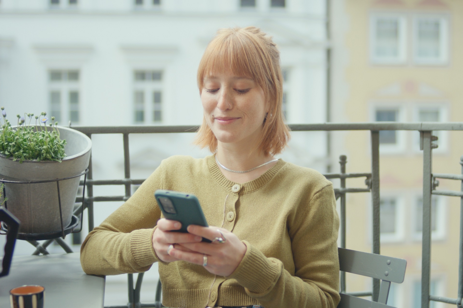 Eine junge Frau sitzt auf dem Balkon und tippt auf ihrem Handy.