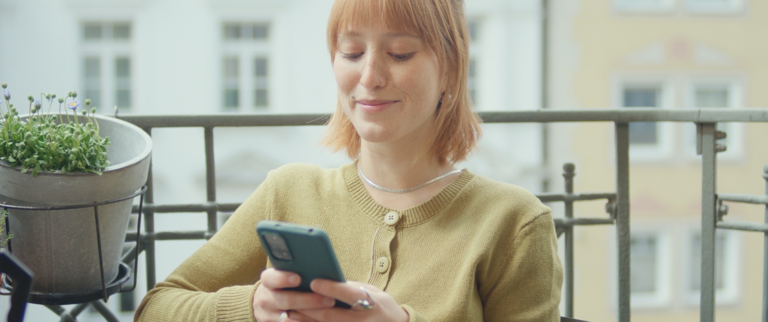 Eine junge Frau sitzt auf dem Balkon und tippt auf ihrem Handy.