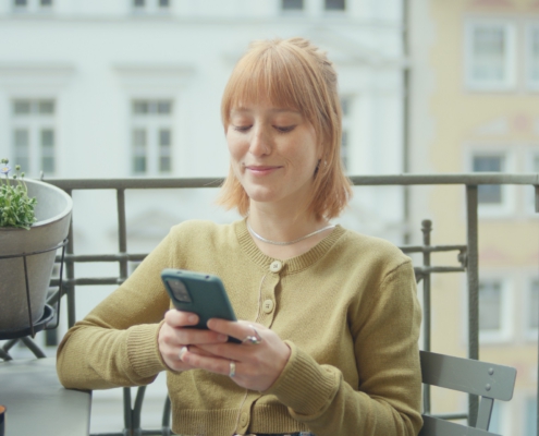 Eine junge Frau sitzt auf dem Balkon und tippt auf ihrem Handy.