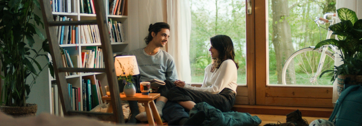 Ein Mann und eine Frau sitzen im Wohnzimmer vor einem Fenster und unterhalten sich. Links von ihnen steht ein großes Bücherregal und ein kleiner Tisch mit einer Lampe.