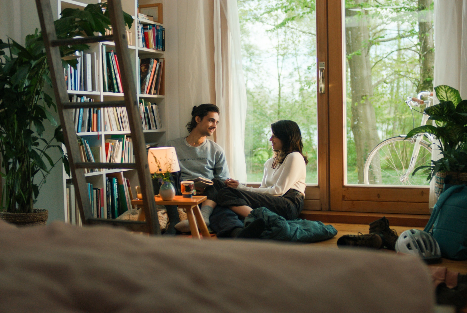 Ein Mann und eine Frau sitzen im Wohnzimmer vor einem Fenster und unterhalten sich. Links von ihnen steht ein großes Bücherregal und ein kleiner Tisch mit einer Lampe. Ein Mann und eine Frau sitzen im Wohnzimmer vor einem Fenster und unterhalten sich. Links von ihnen steht ein großes Bücherregal und ein kleiner Tisch mit einer Lampe.