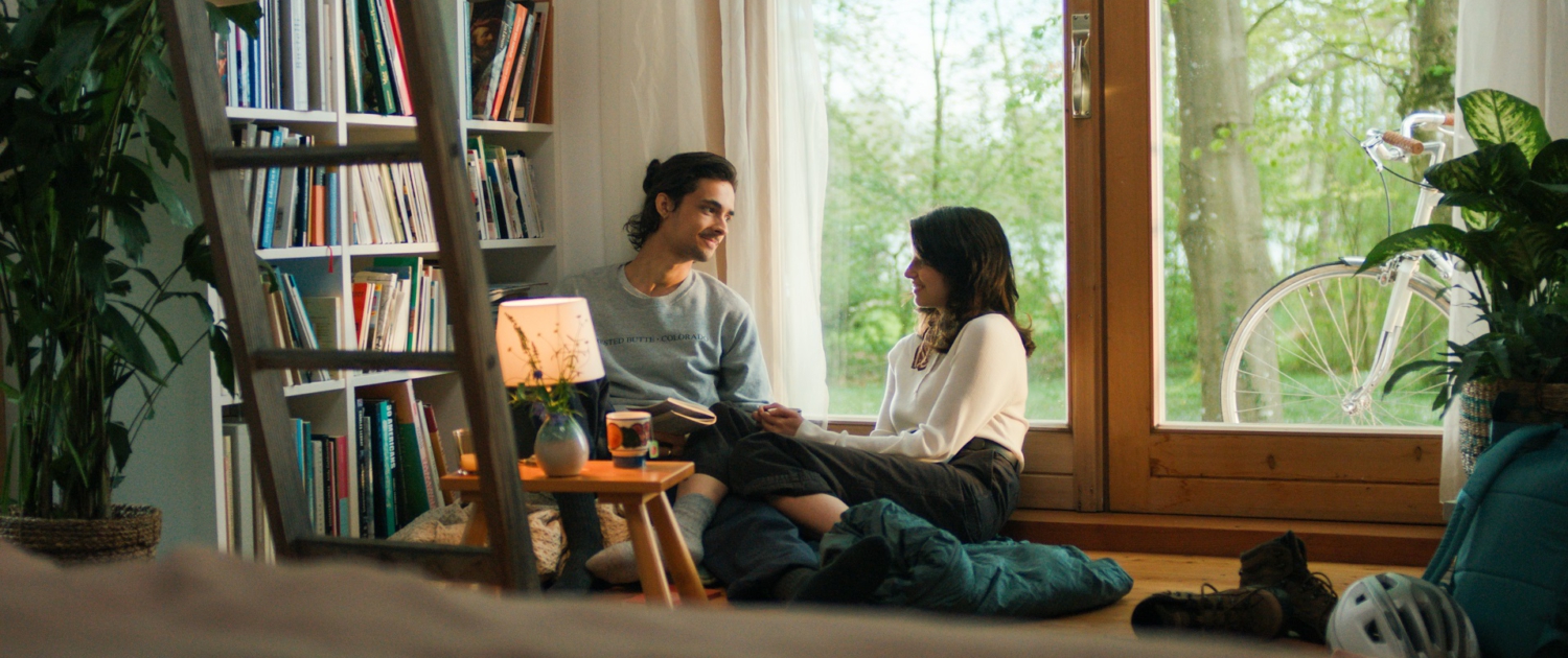 Ein Mann und eine Frau sitzen im Wohnzimmer vor einem Fenster und unterhalten sich. Links von ihnen steht ein großes Bücherregal und ein kleiner Tisch mit einer Lampe.