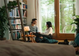Ein Mann und eine Frau sitzen im Wohnzimmer vor einem Fenster und unterhalten sich. Links von ihnen steht ein großes Bücherregal und ein kleiner Tisch mit einer Lampe.