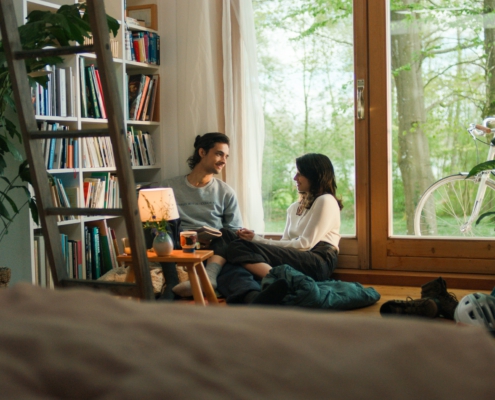 Ein Mann und eine Frau sitzen im Wohnzimmer vor einem Fenster und unterhalten sich. Links von ihnen steht ein großes Bücherregal und ein kleiner Tisch mit einer Lampe.