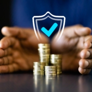 Zwei Hände umschließen einen Stapel Münzen. Darüber ist ein Schild mit einem blauen Hacken projiziert, dass die Sicherheit des Geldes symbolisiert.