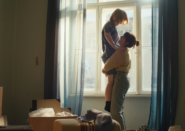 Zwei junge Frauen stehen in einer Wohnung mit Umzugskartons vor dem Fenster. Die eine hebt die andere voller Freude hoch.