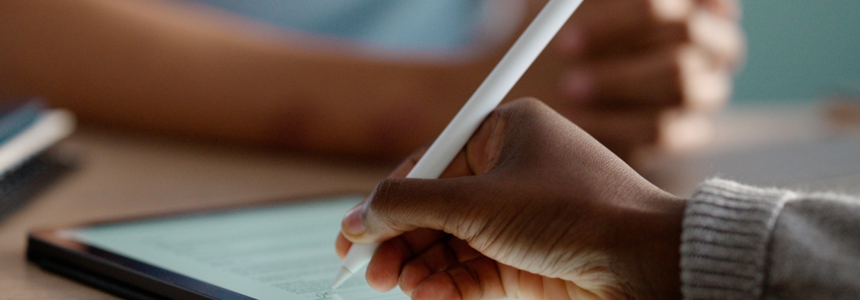 Im Fokus liegt ein Tablet auf einem Tisch. Eine Hand hält einen Stift und unterschreibt ein digitales Formular auf dem Bildschirm.