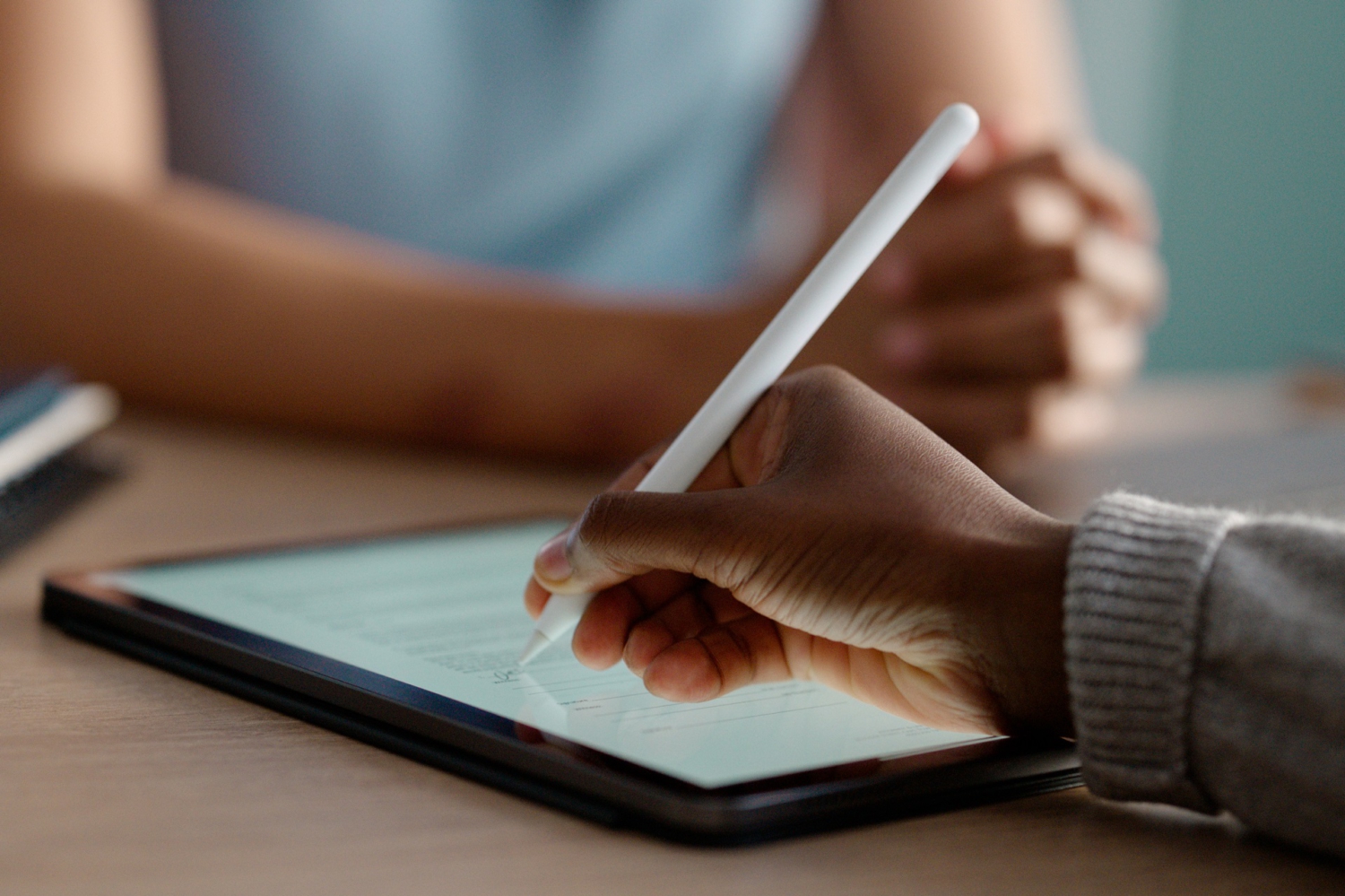 Im Fokus liegt ein Tablet auf einem Tisch. Eine Hand hält einen Stift und unterschreibt ein digitales Formular auf dem Bildschirm.