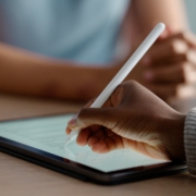 Im Fokus liegt ein Tablet auf einem Tisch. Eine Hand hält einen Stift und unterschreibt ein digitales Formular auf dem Bildschirm.