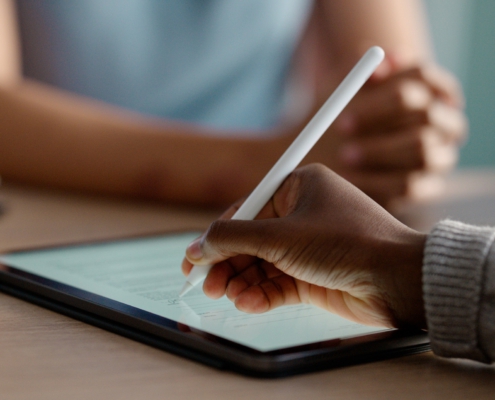 Im Fokus liegt ein Tablet auf einem Tisch. Eine Hand hält einen Stift und unterschreibt ein digitales Formular auf dem Bildschirm.