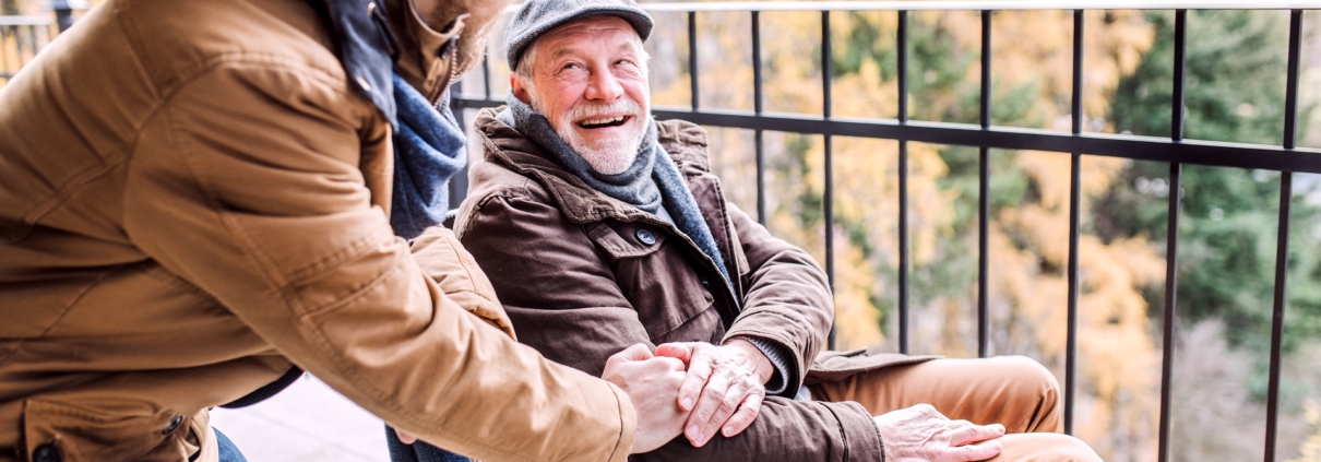 Ein junger Mann steht seitlich, leicht nach vornge gebeugt neben einem älteren Herren im Rollstuhl. Sie lächeln sich an. Der Ältere hat dem Jüngeren seine Hand auf dessen Hand gelegt.