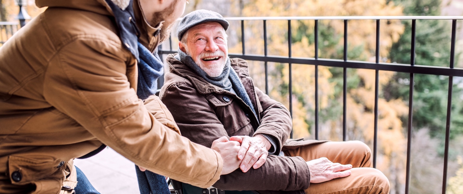 Ein junger Mann steht seitlich, leicht nach vornge gebeugt neben einem älteren Herren im Rollstuhl. Sie lächeln sich an. Der Ältere hat dem Jüngeren seine Hand auf dessen Hand gelegt. Ein junger Mann steht seitlich, leicht nach vornge gebeugt neben einem älteren Herren im Rollstuhl. Sie lächeln sich an. Der Ältere hat dem Jüngeren seine Hand auf dessen Hand gelegt.