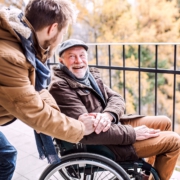 Ein junger Mann steht seitlich, leicht nach vornge gebeugt neben einem älteren Herren im Rollstuhl. Sie lächeln sich an. Der Ältere hat dem Jüngeren seine Hand auf dessen Hand gelegt.