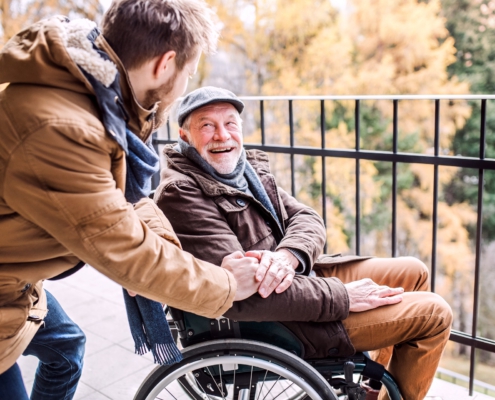 Ein junger Mann steht seitlich, leicht nach vornge gebeugt neben einem älteren Herren im Rollstuhl. Sie lächeln sich an. Der Ältere hat dem Jüngeren seine Hand auf dessen Hand gelegt.