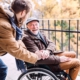 Ein junger Mann steht seitlich, leicht nach vornge gebeugt neben einem älteren Herren im Rollstuhl. Sie lächeln sich an. Der Ältere hat dem Jüngeren seine Hand auf dessen Hand gelegt.