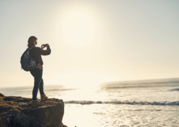 Links im Bild steht eine Frau an einer Klippe und fotografiert auf das offene Meer hinaus. Im Hintergrund scheint die Abendsonne.