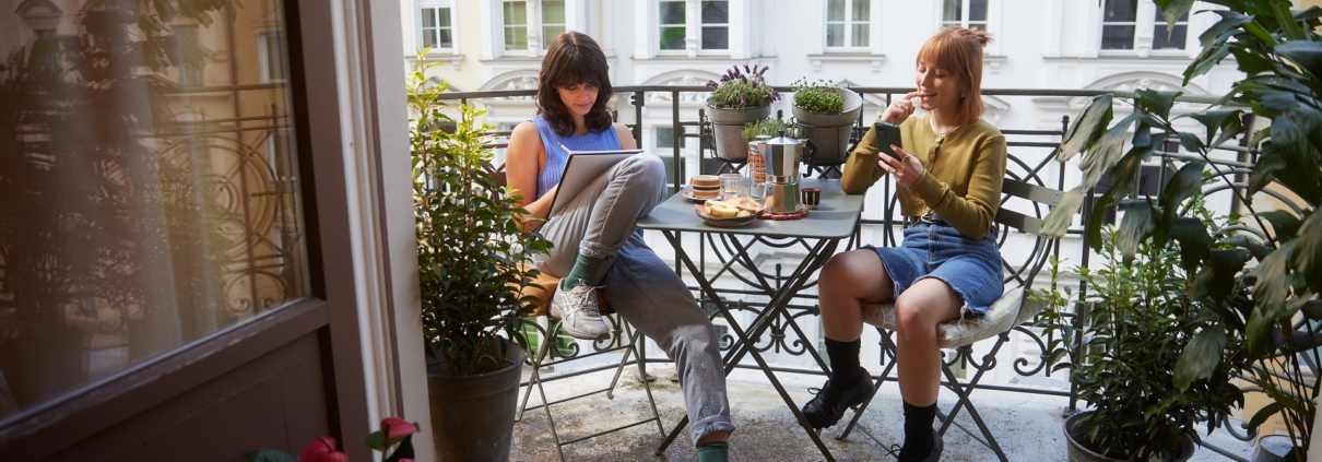 Zwei junge Frauen sitzen sich bei schönem Wetter auf einem Balkon an einem mit Snacks und Kaffee gedecktem Tisch gegenüber. Links, mit braunen Haaren, macht sich notizen. Rechts, mit roten Haaren, schaut in ihr Handy.