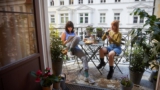 Zwei junge Frauen sitzen sich bei schönem Wetter auf einem Balkon an einem mit Snacks und Kaffee gedecktem Tisch gegenüber. Links, mit braunen Haaren, macht sich notizen. Rechts, mit roten Haaren, schaut in ihr Handy.