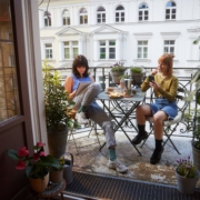 Zwei junge Frauen sitzen sich bei schönem Wetter auf einem Balkon an einem mit Snacks und Kaffee gedecktem Tisch gegenüber. Links, mit braunen Haaren, macht sich notizen. Rechts, mit roten Haaren, schaut in ihr Handy.