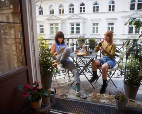 Zwei junge Frauen sitzen sich bei schönem Wetter auf einem Balkon an einem mit Snacks und Kaffee gedecktem Tisch gegenüber. Links, mit braunen Haaren, macht sich notizen. Rechts, mit roten Haaren, schaut in ihr Handy.