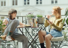 Zwei junge Frauen sitzen sich bei schönem Wetter auf einem Balkon an einem mit Snacks und Kaffee gedecktem Tisch gegenüber. Sie unterhalten sich. Zwei junge Frauen sitzen sich bei schönem Wetter auf einem Balkon an einem mit Snacks und Kaffee gedecktem Tisch gegenüber. Sie unterhalten sich.