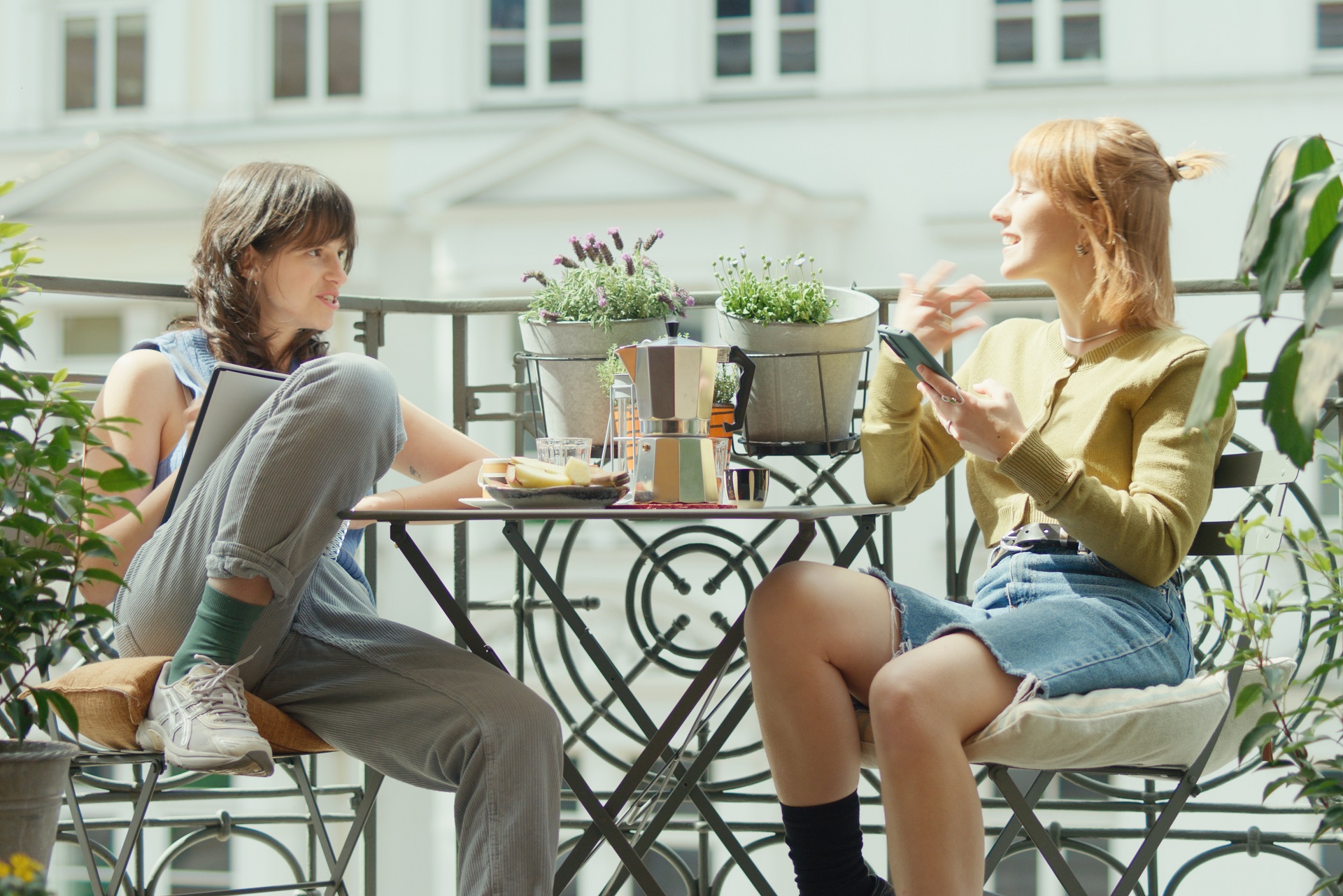 Zwei junge Frauen sitzen sich bei schönem Wetter auf einem Balkon an einem mit Snacks und Kaffee gedecktem Tisch gegenüber. Sie unterhalten sich.