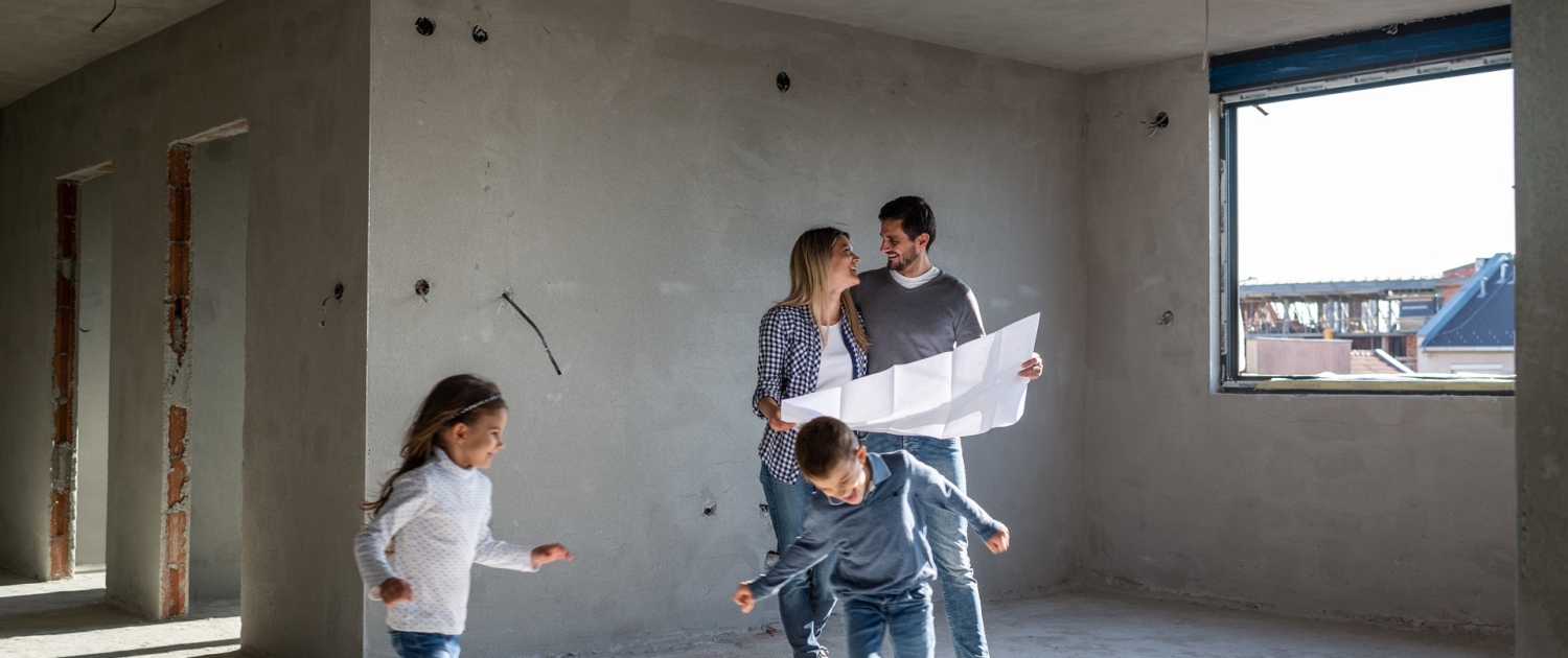 In der Mitte steht ein Pärchen in einem Rohbau. Es fehlen noch Fenster und Türen. Um sie herum rennen zwei Kinder, ein Junge und ein Mädchen. Die Eltern halten einen Plan in der Hand und lächeln sich an.