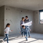 In der Mitte steht ein Pärchen in einem Rohbau. Es fehlen noch Fenster und Türen. Um sie herum rennen zwei Kinder, ein Junge und ein Mädchen. Die Eltern halten einen Plan in der Hand und lächeln sich an.