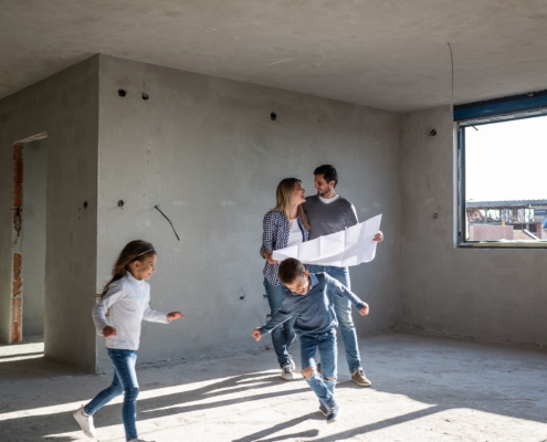 In der Mitte steht ein Pärchen in einem Rohbau. Es fehlen noch Fenster und Türen. Um sie herum rennen zwei Kinder, ein Junge und ein Mädchen. Die Eltern halten einen Plan in der Hand und lächeln sich an.