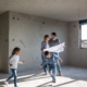 In der Mitte steht ein Pärchen in einem Rohbau. Es fehlen noch Fenster und Türen. Um sie herum rennen zwei Kinder, ein Junge und ein Mädchen. Die Eltern halten einen Plan in der Hand und lächeln sich an.
