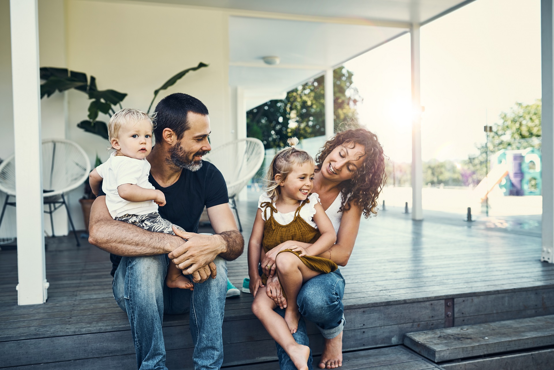 aEin Mann und eine Frau sitzen auf einer Veranda vor einem Haus. Er hat einen kleinen jungen auf dem Schoß, sie ein kleines Mädchen. Die Familie ist fröhlich und lächelt.