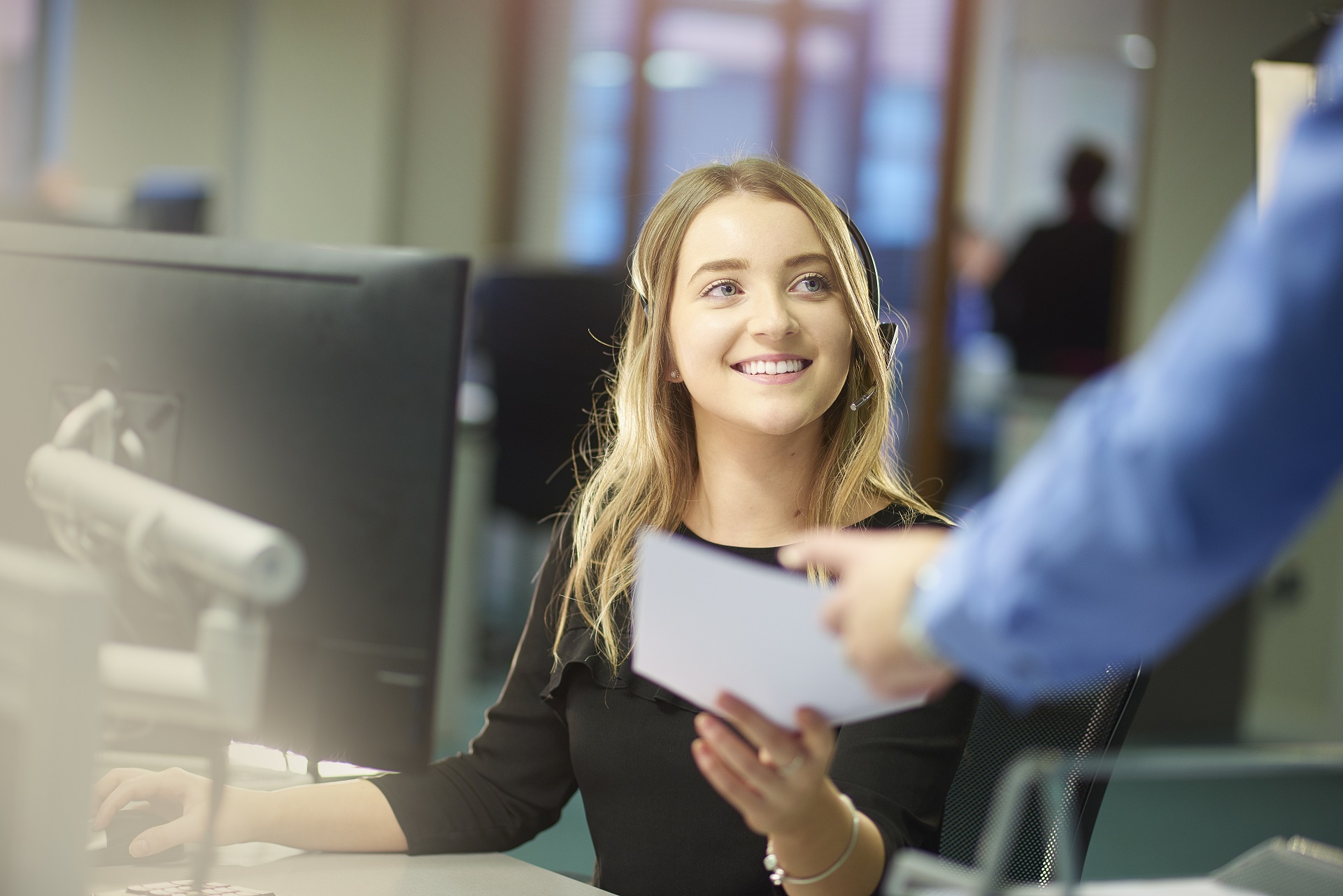 Eine blonde Frau sitz an einem Büroschreibtisch. Sie Trägt ein Headset. Im Vordergrund sieht man seitlich verschommen eine Hand, die der Frau was gibt. Sie nimmt es lächeln and. Eine blonde Frau sitz an einem Büroschreibtisch. Sie Trägt ein Headset. Im Vordergrund sieht man seitlich verschommen eine Hand, die der Frau was gibt. Sie nimmt es lächeln and.