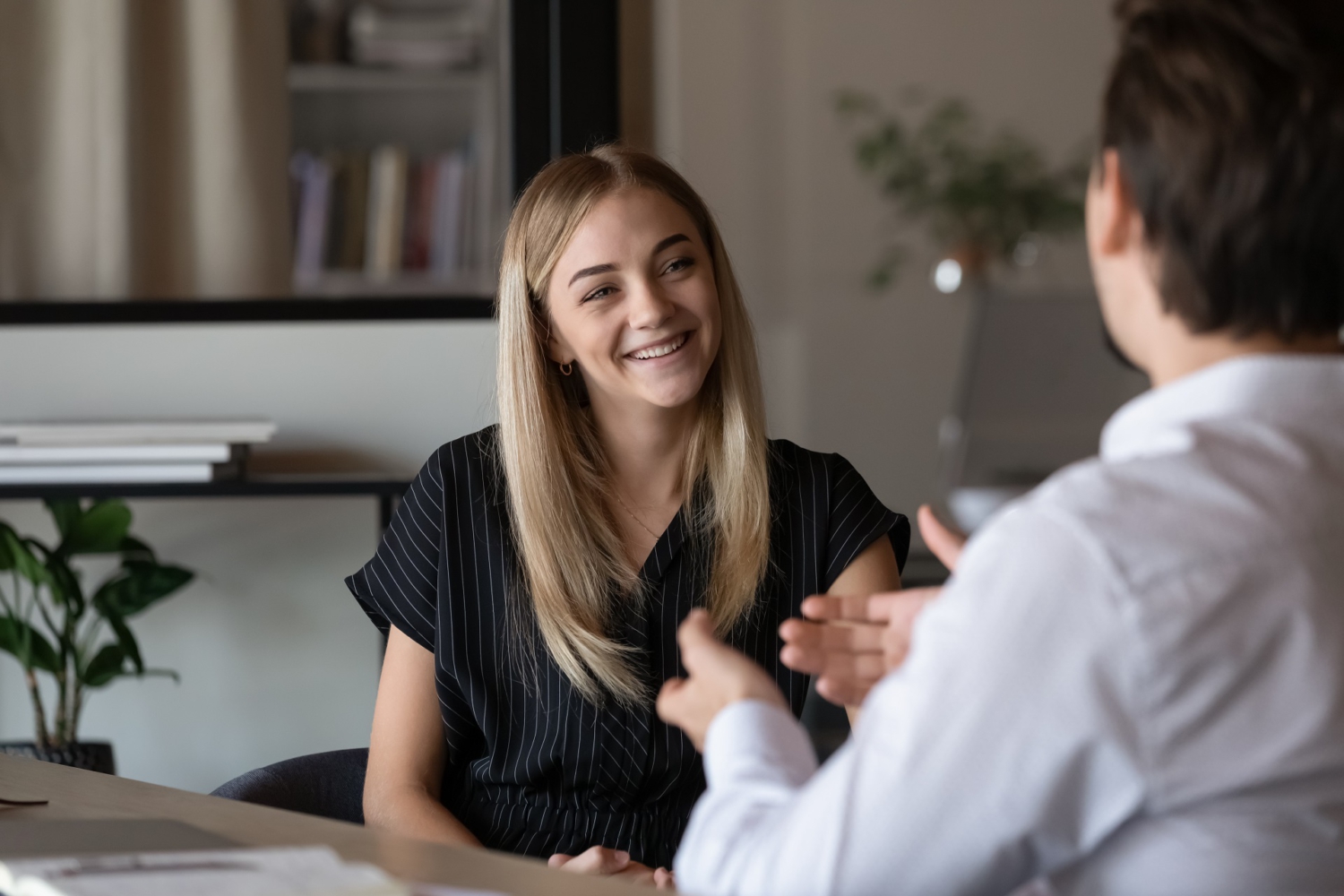 In der Mitte sitzt eine Frau seitlich an einem Tisch. Ihr gegenüber, mit dem Rücken zum Bild, sitz ein Mann. Die beiden unterhalten sich in einem Büro. Er gestikuliert, sie lächelt. In der Mitte sitzt eine Frau seitlich an einem Tisch. Ihr gegenüber, mit dem Rücken zum Bild, sitz ein Mann. Die beiden unterhalten sich in einem Büro. Er gestikuliert, sie lächelt.