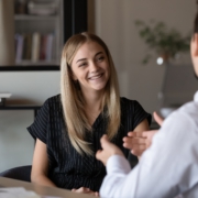 In der Mitte sitzt eine Frau seitlich an einem Tisch. Ihr gegenüber, mit dem Rücken zum Bild, sitz ein Mann. Die beiden unterhalten sich in einem Büro. Er gestikuliert, sie lächelt.