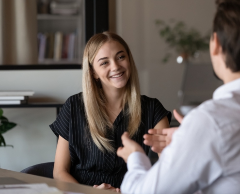 In der Mitte sitzt eine Frau seitlich an einem Tisch. Ihr gegenüber, mit dem Rücken zum Bild, sitz ein Mann. Die beiden unterhalten sich in einem Büro. Er gestikuliert, sie lächelt.