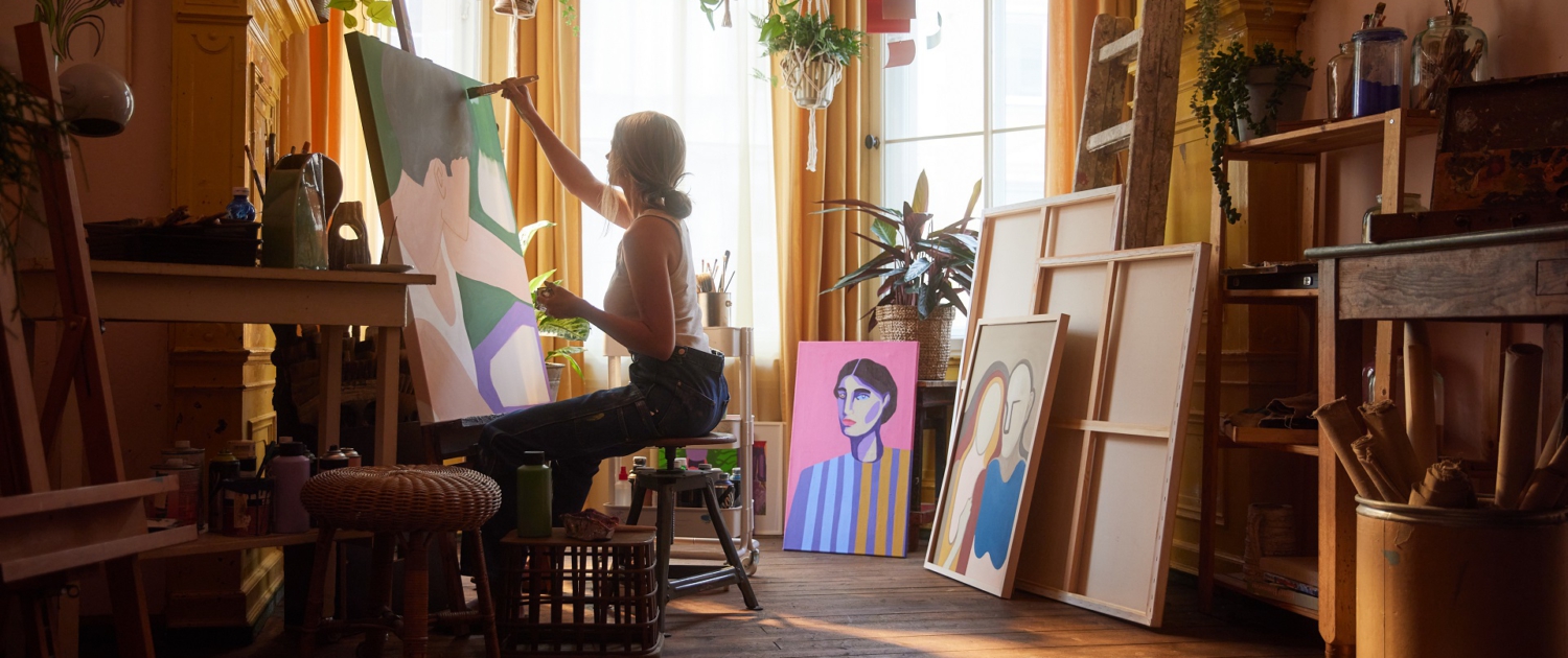 Eine junge Künstlerin sitzt vor einer Staffelei und malt in ihrem Atelier ein Bild. Darauf ist eine Person mit kurzen Haaren und weißen Top seitlich von oben zu sehen. Eine junge Künstlerin sitzt vor einer Staffelei und malt in ihrem Atelier ein Bild. Darauf ist eine Person mit kurzen Haaren und weißen Top seitlich von oben zu sehen.