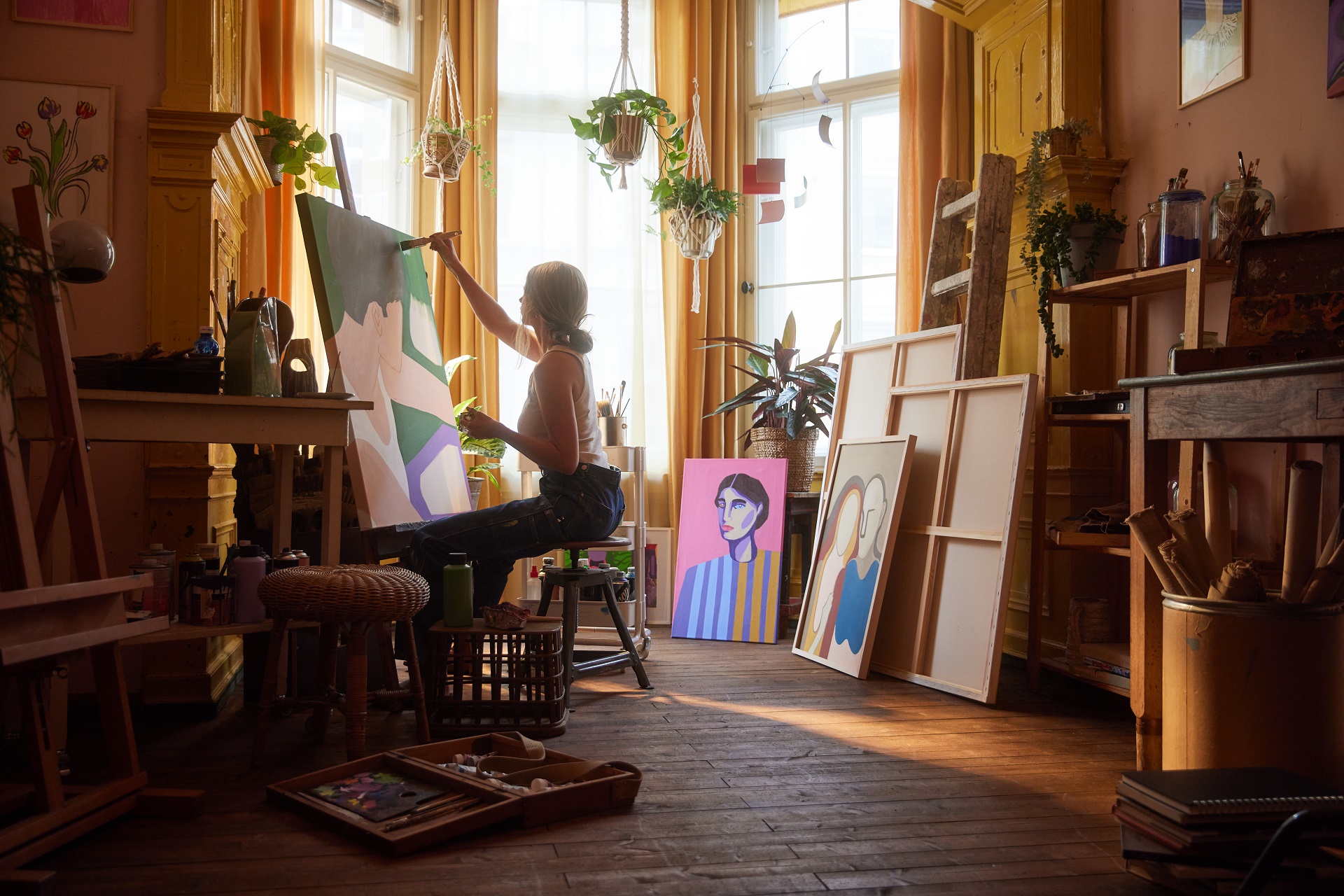 Eine junge Künstlerin sitzt vor einer Staffelei und malt in ihrem Atelier ein Bild. Darauf ist eine Person mit kurzen Haaren und weißen Top seitlich von oben zu sehen.