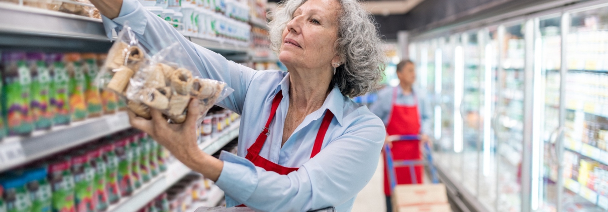 Eine Frau mittleren Alters steht auf einer Leiter und räumt Kekse in das oberste Fach eines Regals ein. Im Hintergrund erkennt man die Kühlregale eines Supermarktes.