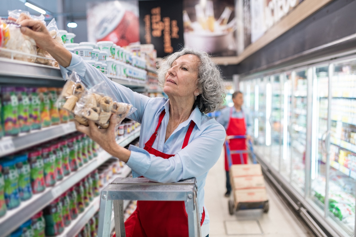 Eine Frau mittleren Alters steht auf einer Leiter und räumt Kekse in das oberste Fach eines Regals ein. Im Hintergrund erkennt man die Kühlregale eines Supermarktes. Eine Frau mittleren Alters steht auf einer Leiter und räumt Kekse in das oberste Fach eines Regals ein. Im Hintergrund erkennt man die Kühlregale eines Supermarktes.