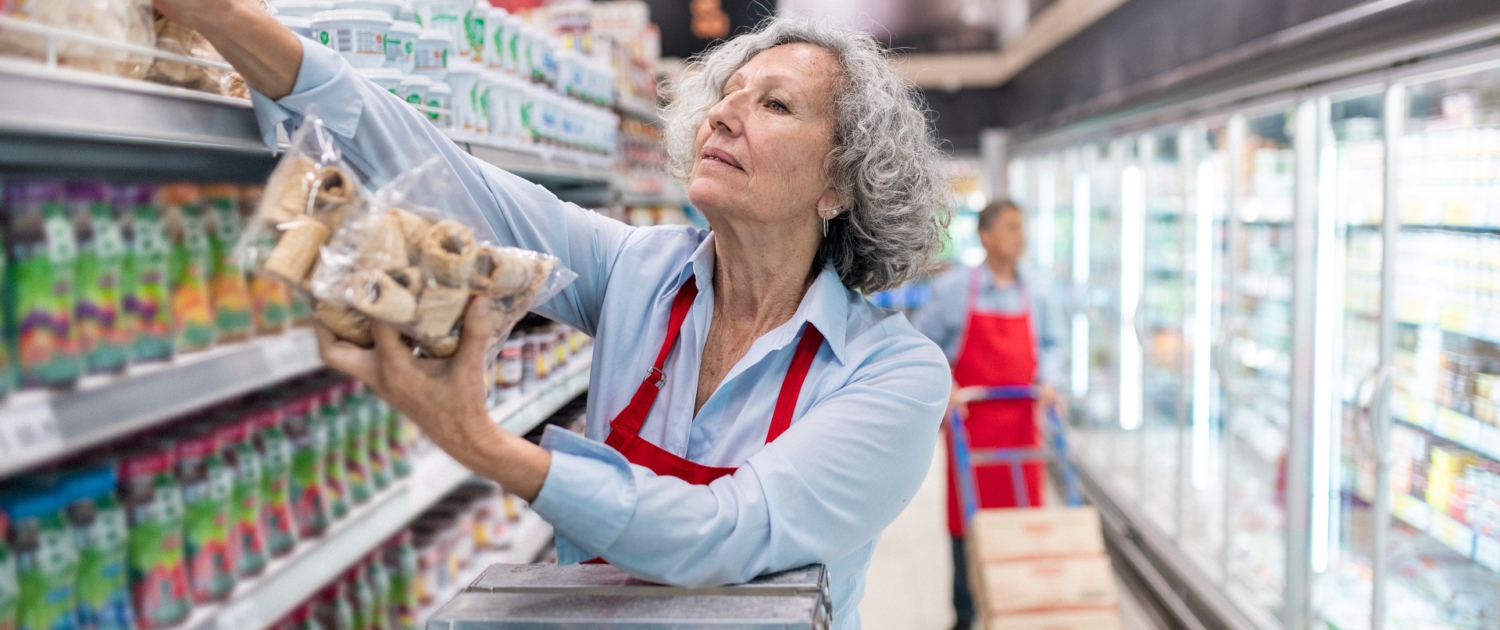 Eine Frau mittleren Alters steht auf einer Leiter und räumt Kekse in das oberste Fach eines Regals ein. Im Hintergrund erkennt man die Kühlregale eines Supermarktes. Eine Frau mittleren Alters steht auf einer Leiter und räumt Kekse in das oberste Fach eines Regals ein. Im Hintergrund erkennt man die Kühlregale eines Supermarktes.