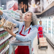 Eine Frau mittleren Alters steht auf einer Leiter und räumt Kekse in das oberste Fach eines Regals ein. Im Hintergrund erkennt man die Kühlregale eines Supermarktes.