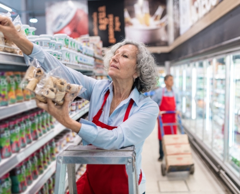 Eine Frau mittleren Alters steht auf einer Leiter und räumt Kekse in das oberste Fach eines Regals ein. Im Hintergrund erkennt man die Kühlregale eines Supermarktes.