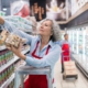 Eine Frau mittleren Alters steht auf einer Leiter und räumt Kekse in das oberste Fach eines Regals ein. Im Hintergrund erkennt man die Kühlregale eines Supermarktes.