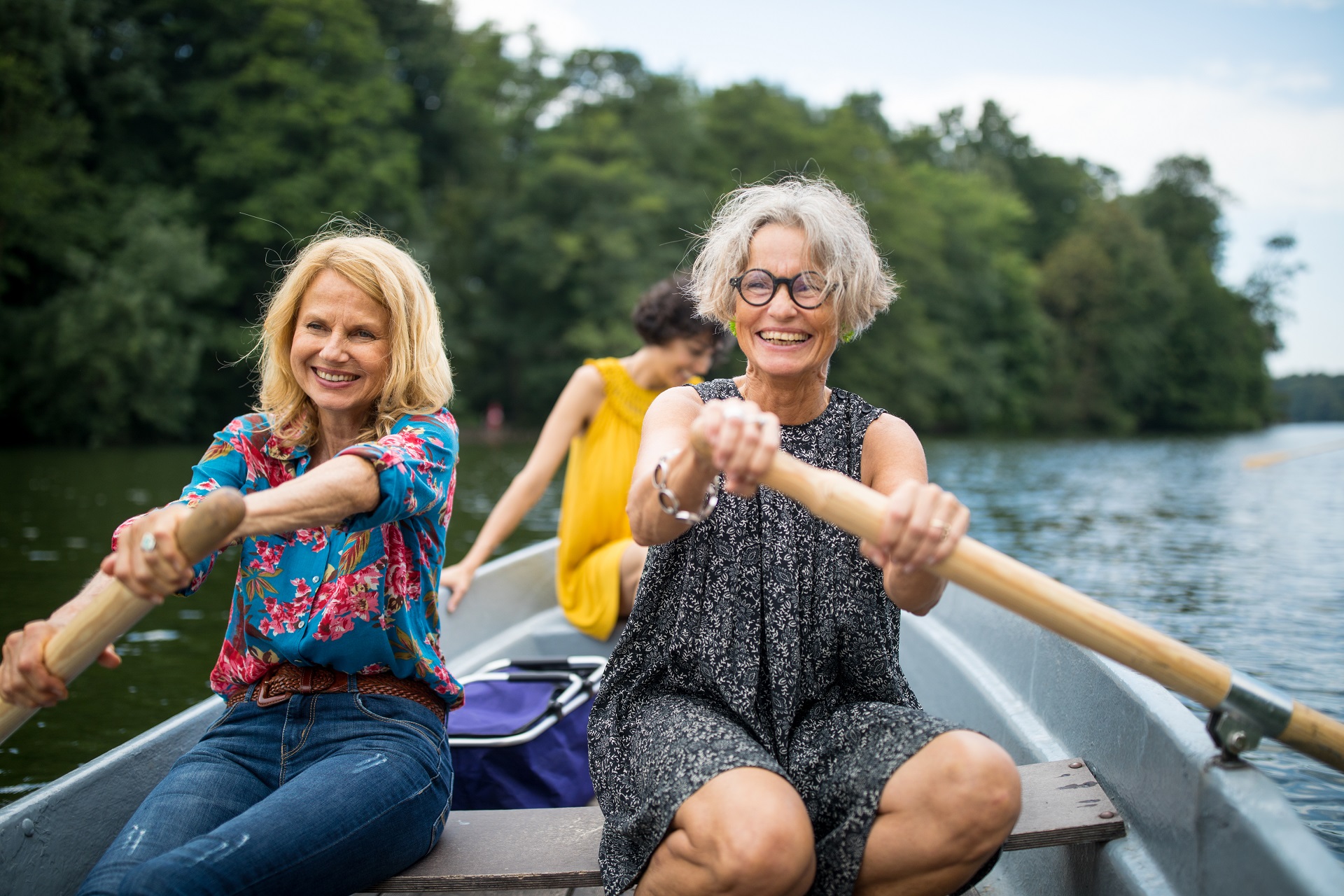Zwei Frauen mittleren Alters sitzen in einem Ruderboot und rudern. Im Hintergrund erkennt man eine jüngere Frau. Sie befindne sich mit dem Ruderbooot auf einem See. Zwei Frauen mittleren Alters sitzen in einem Ruderboot und rudern. Im Hintergrund erkennt man eine jüngere Frau. Sie befindne sich mit dem Ruderbooot auf einem See.