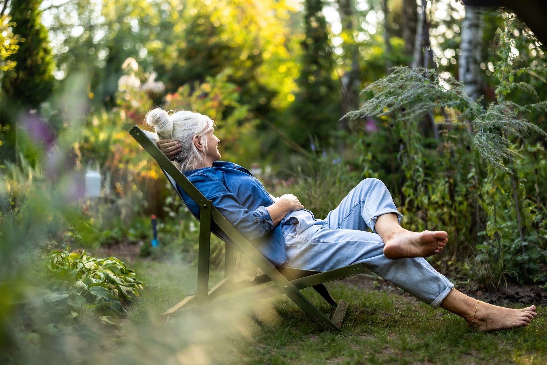 Eine ältere aber noch sehr fitte Frau sitzt in einem Liegestuhl mitten in einem grünen Garten. Sie blickt verträumt in die Ferne.