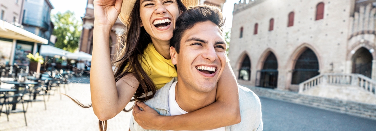 aIm Vordergrund ist ein Pärchen. Er hat sie Huckepack. Beide lachen. Im Hintergrund ist die Szenerie einer kleinen alten Stadt mit Vorplatz zu erkennen.