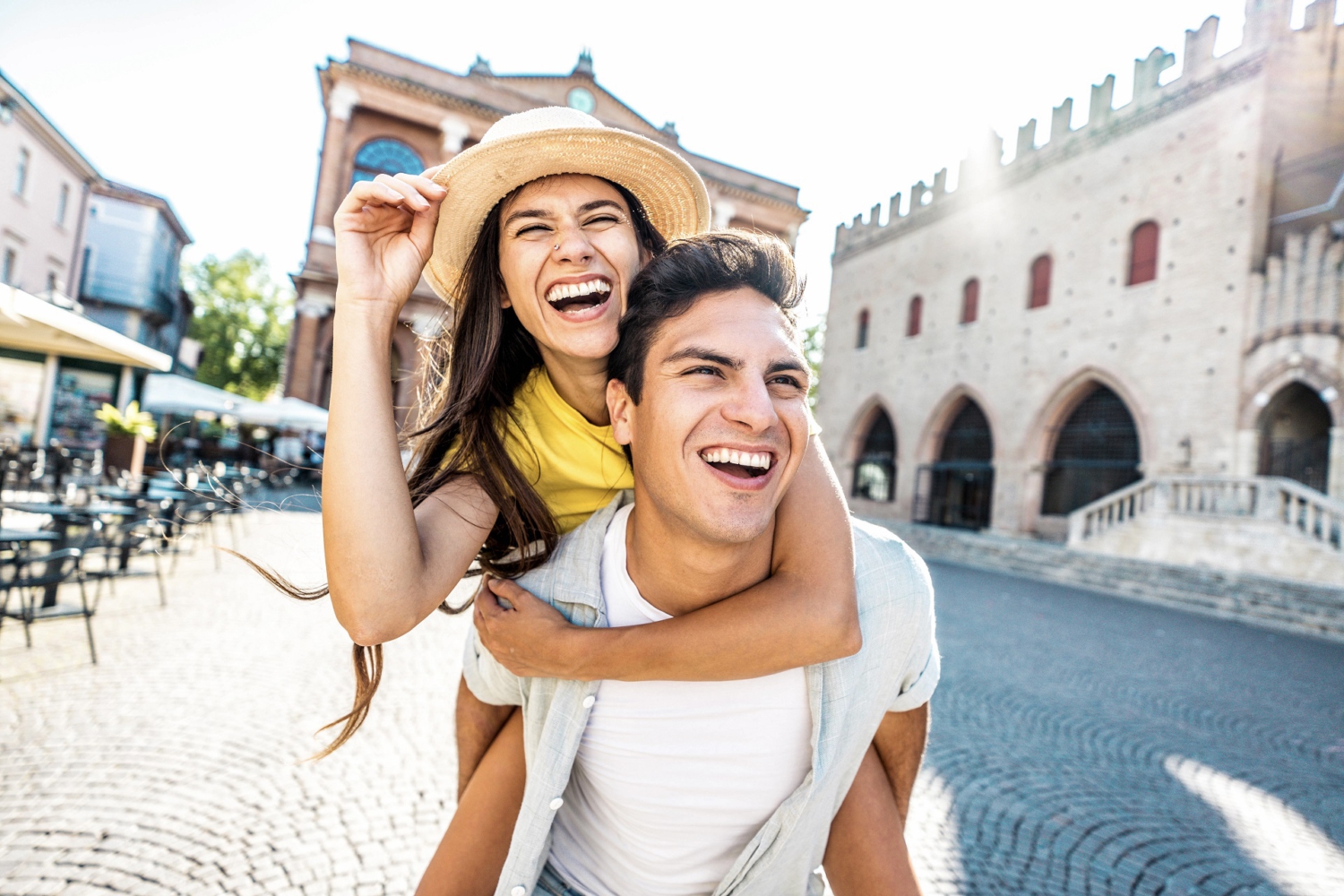 Im Vordergrund ist ein Pärchen. Er hat sie Huckepack. Beide lachen. Im Hintergrund ist die Szenerie einer kleinen alten Stadt mit Vorplatz zu erkennen. aIm Vordergrund ist ein Pärchen. Er hat sie Huckepack. Beide lachen. Im Hintergrund ist die Szenerie einer kleinen alten Stadt mit Vorplatz zu erkennen.