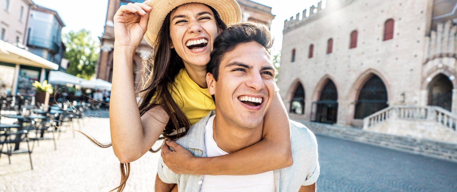 Im Vordergrund ist ein Pärchen. Er hat sie Huckepack. Beide lachen. Im Hintergrund ist die Szenerie einer kleinen alten Stadt mit Vorplatz zu erkennen. aIm Vordergrund ist ein Pärchen. Er hat sie Huckepack. Beide lachen. Im Hintergrund ist die Szenerie einer kleinen alten Stadt mit Vorplatz zu erkennen.