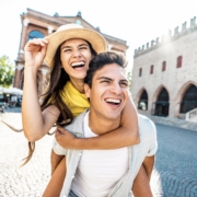 aIm Vordergrund ist ein Pärchen. Er hat sie Huckepack. Beide lachen. Im Hintergrund ist die Szenerie einer kleinen alten Stadt mit Vorplatz zu erkennen.